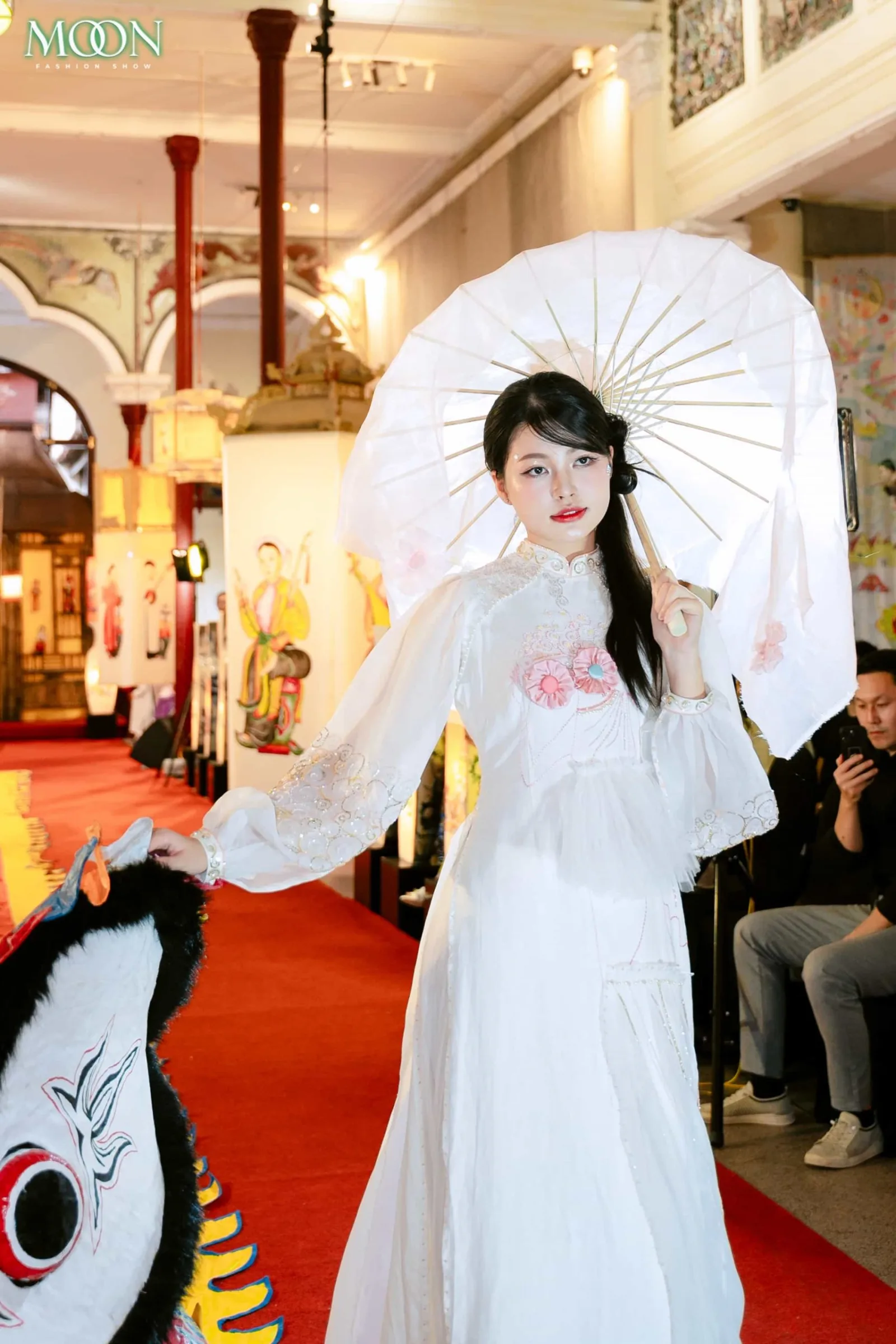 Model in white gown with floral details and parasol walking the runway at Moon Fashion Show 2025 event.