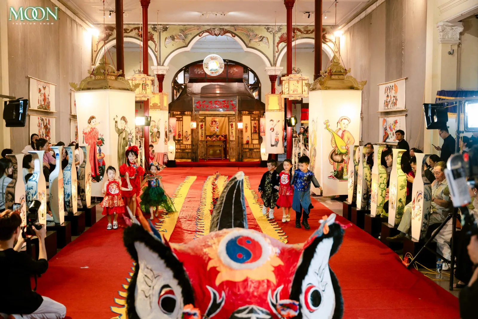 Children in colorful outfits walk the red runway at Moon Fashion Show 2025, surrounded by traditional art displays.