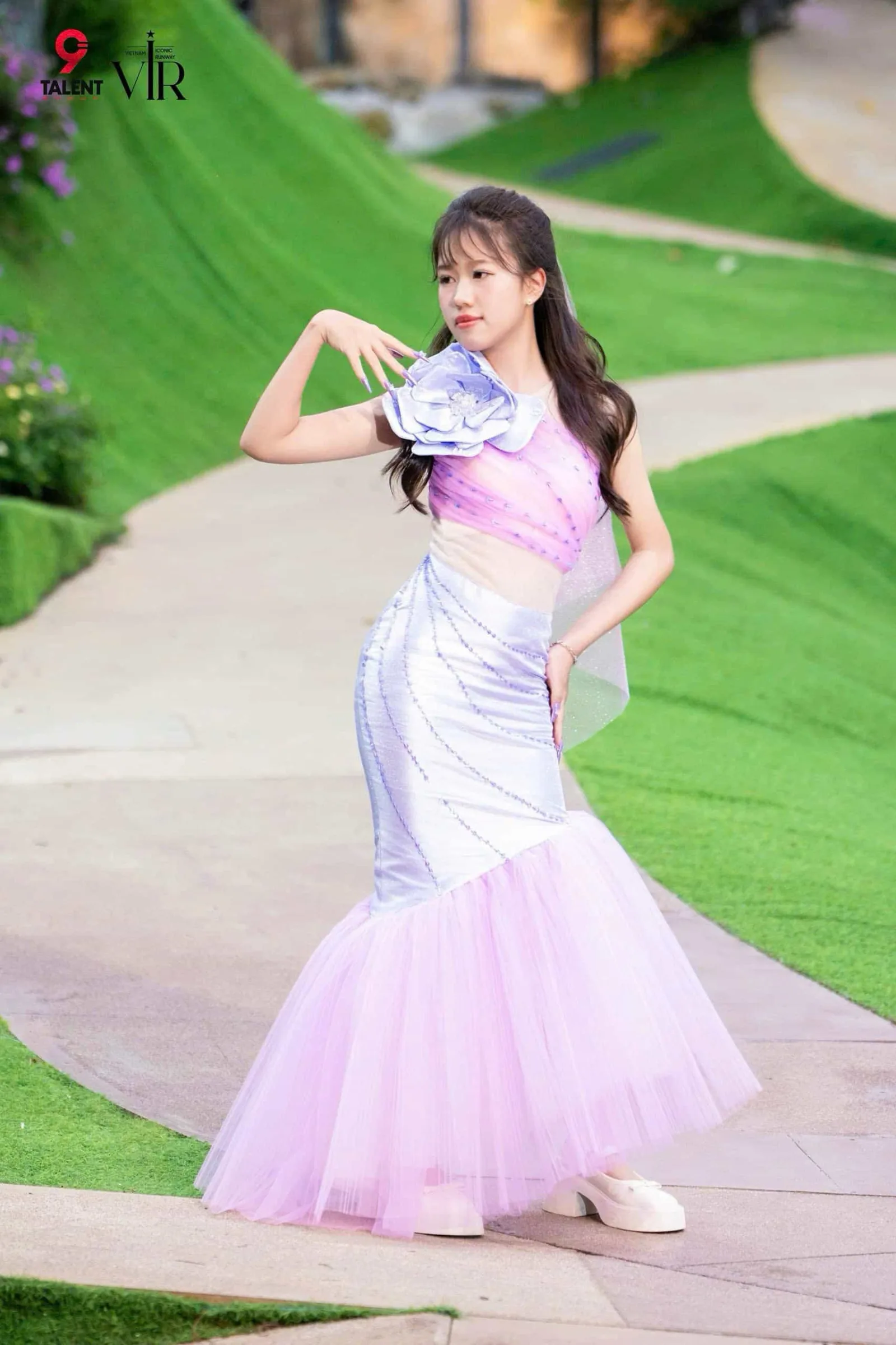 Trần Ngọc Ánh poses gracefully in a pastel mermaid-style dress with floral details on a scenic outdoor pathway.