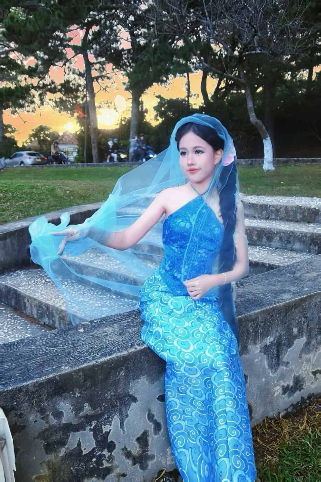 Trần Ngọc Ánh in a flowing blue dress and veil poses gracefully outdoors with a scenic sunset background.